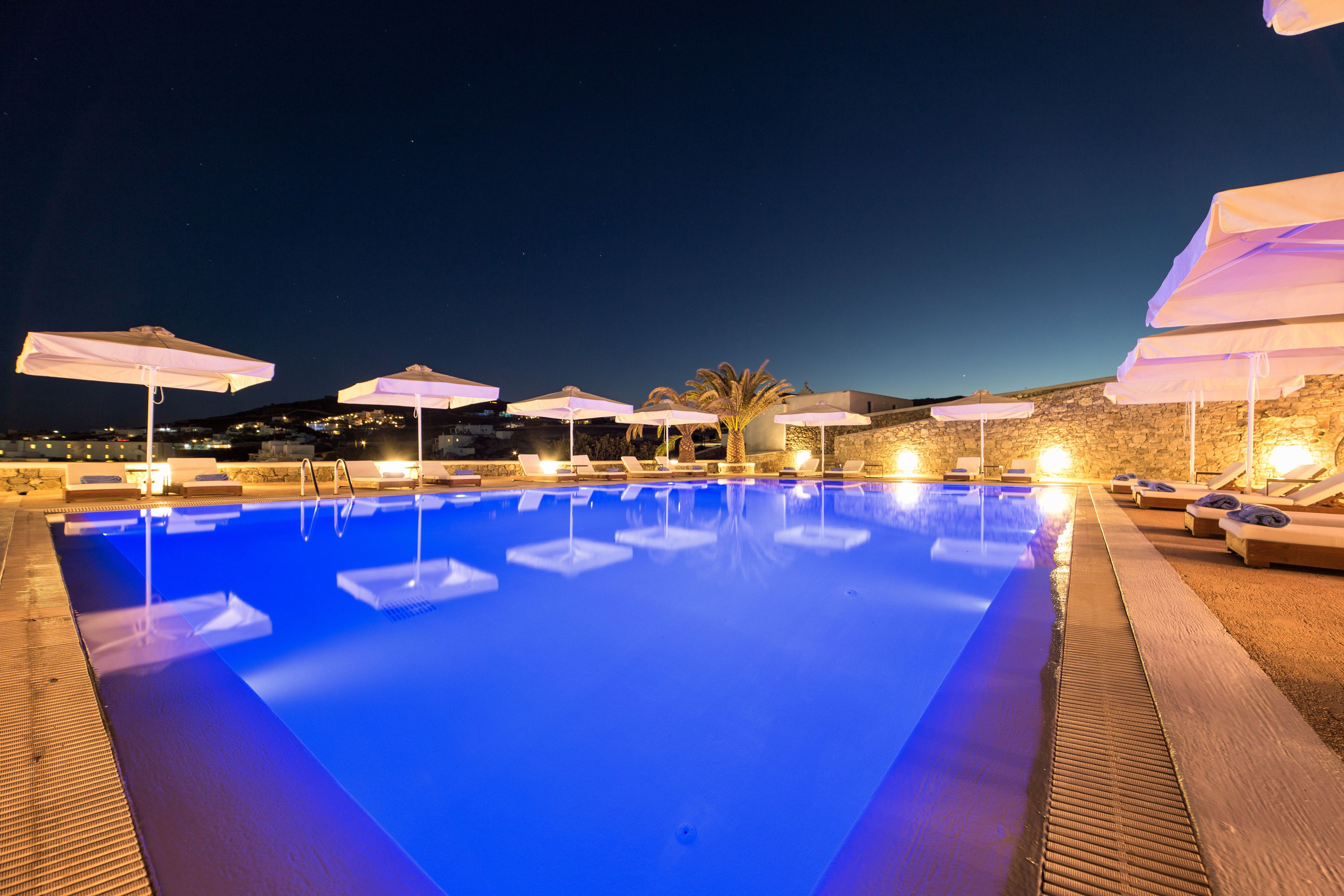 Une piscine nocturne aux eaux bleues illuminées, entourée de chaises longues et de parasols, le tout se détachant sur un décor de bâtiments en pierre et d’un ciel étoilé.