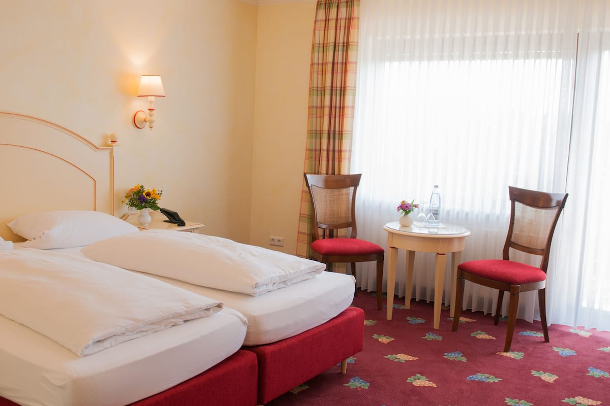 Hotelkamer met een tweepersoonsbed, twee stoelen, een klein tafeltje, een lamp en een raam met gordijnen.