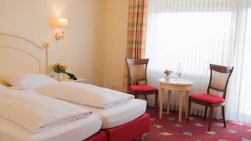 Una habitación de hotel con cama doble, dos sillas, una mesita, lámpara y ventana con cortinas.