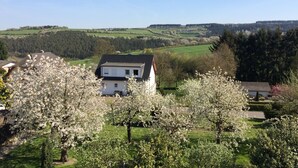 Een huis met een witte gevel en donker dak, omgeven door bloeiende bomen en een groen landschap.