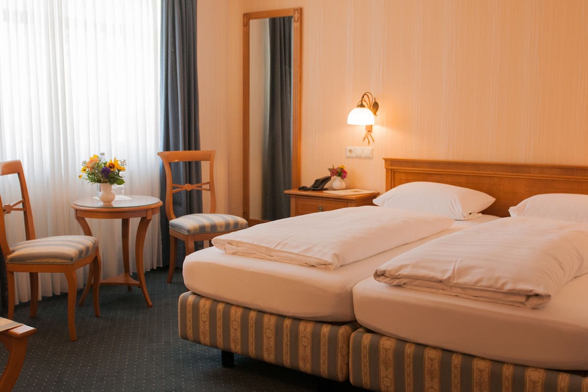 Een hotelkamer met twee bedden, een klein rond tafeltje met een bloevas, een stoel, een nachtkastje met een lamp en een spiegel aan de muur.