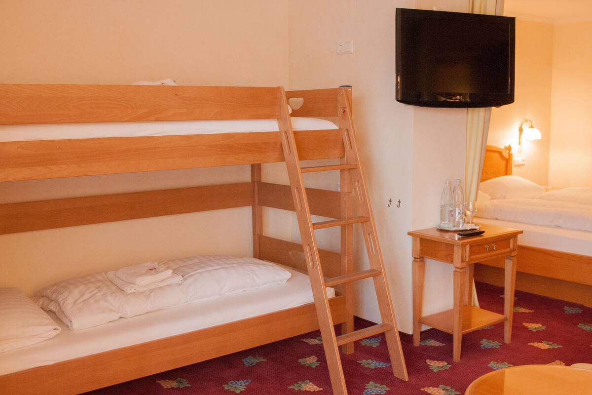 Een stapelbed met een houten trapje, een nachtkastje met een afstandsbediening, en een aan de muur bevestigde televisie.