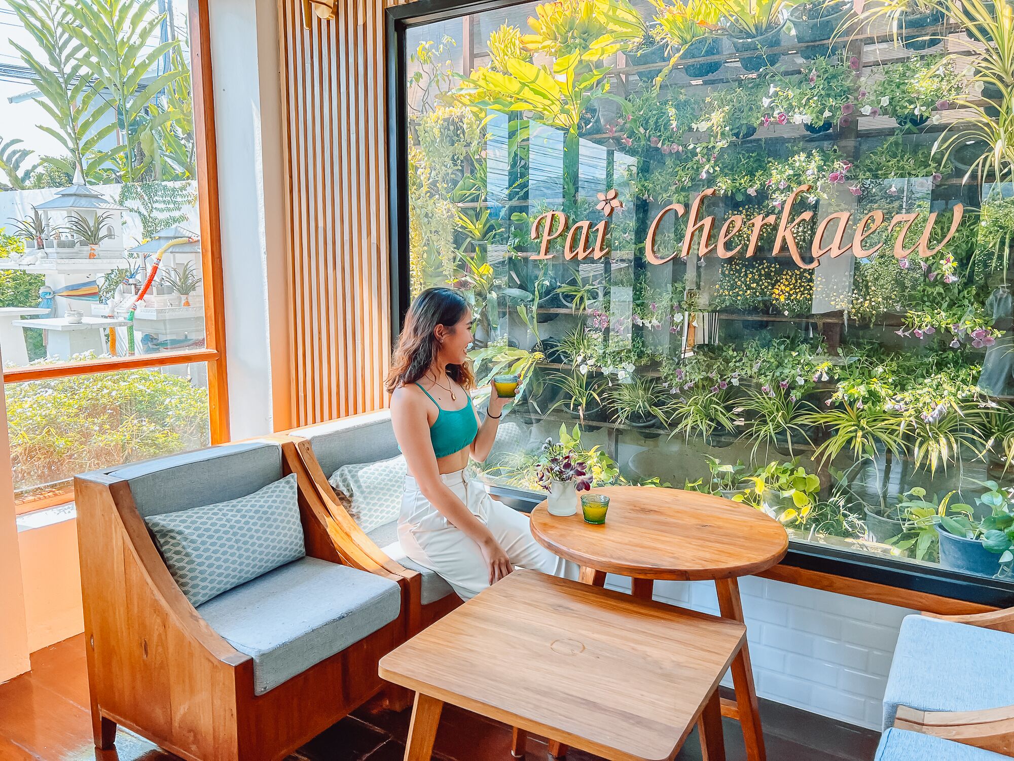 A woman sitting by a window with a view of a lush indoor garden and the words "Pai Cherkaew" on the glass.