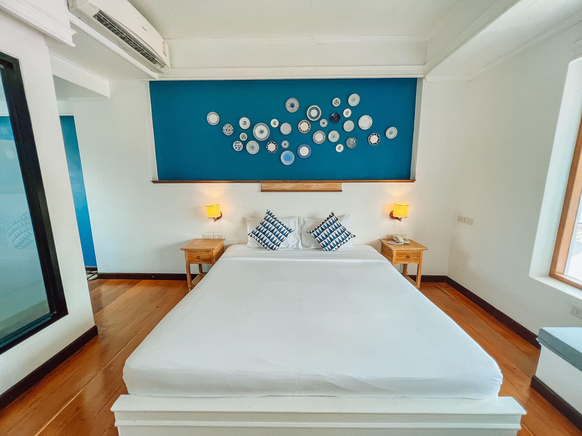A hotel room with a large bed, two bedside tables with lamps, a wall-mounted plate collection, and a large window.