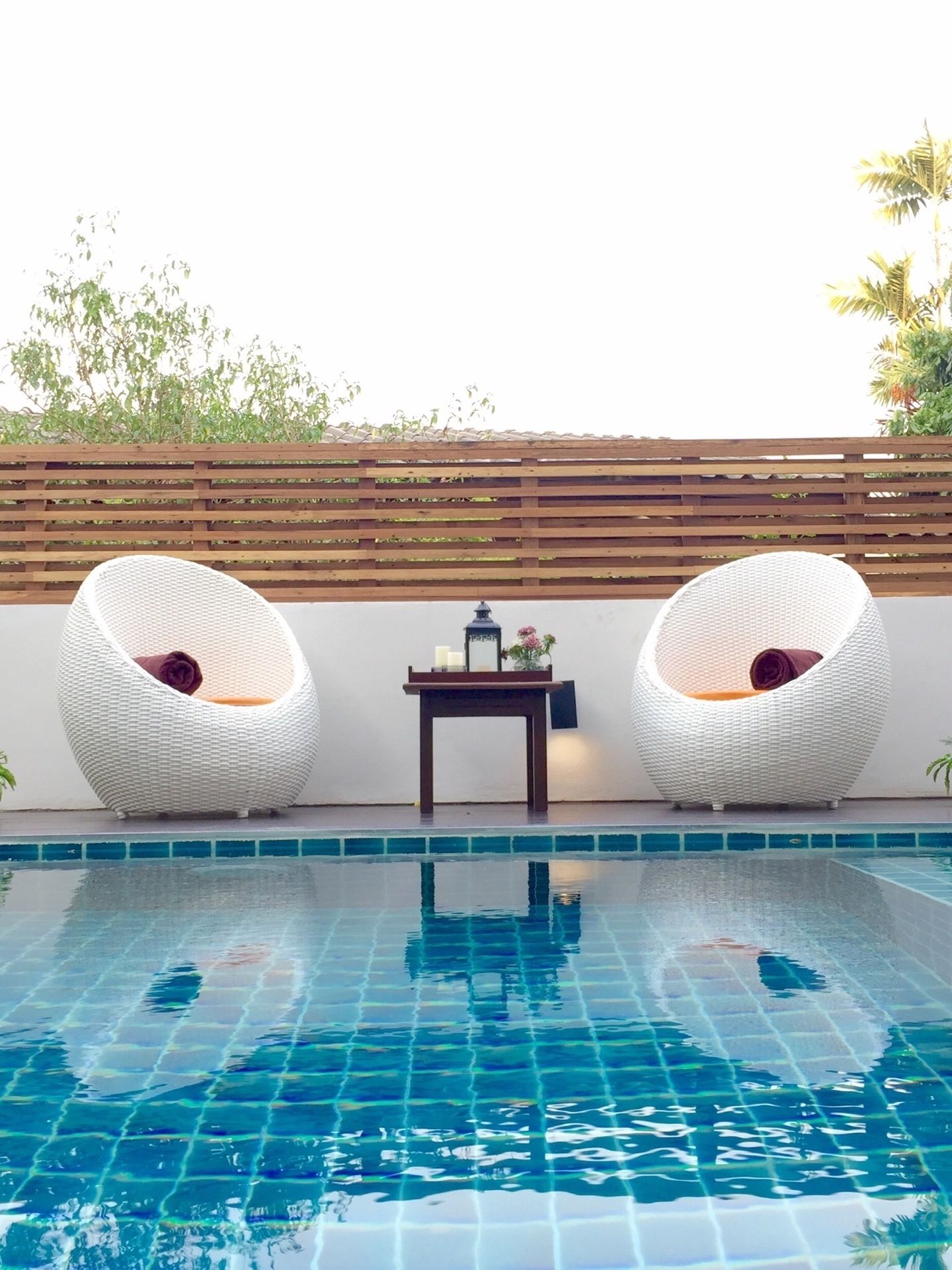 A swimming pool with two white wicker chairs and a small table with a lamp and flowers.