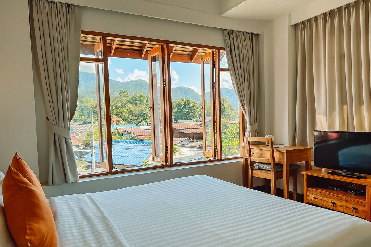Ein Hotelzimmer mit Bett, Schreibtisch mit Fernseher und Blick auf eine gebirgige Landschaft durch große Fenster.