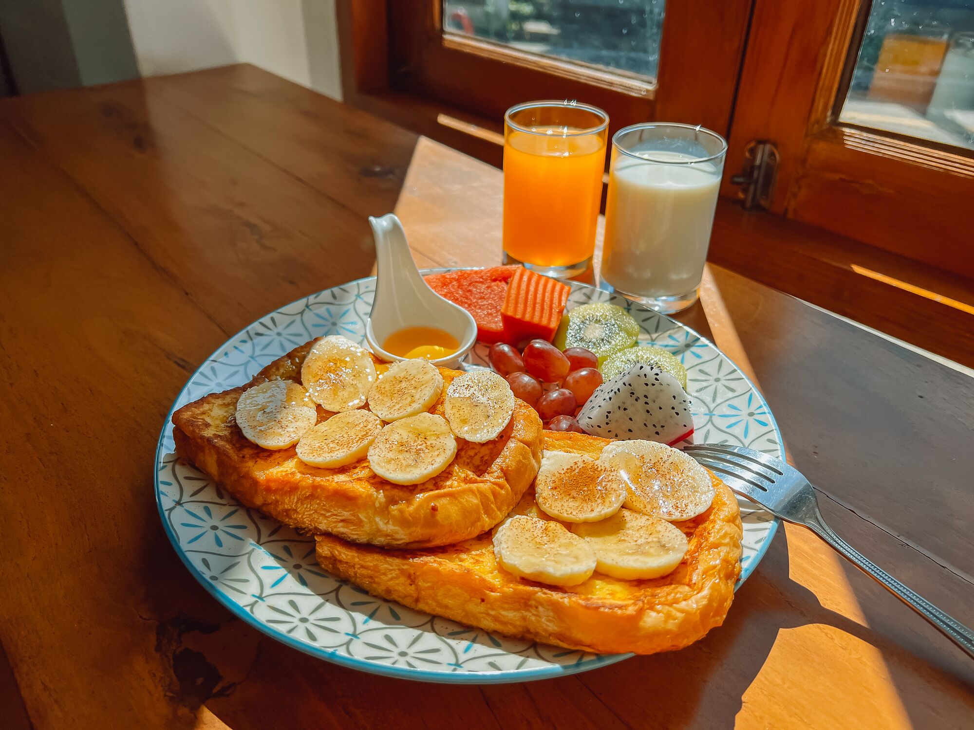 A breakfast plate with a French toast topped with banana slices and a small bowl of syrup, accompanied by a variety of fresh fruits and two glasses of orange juice.