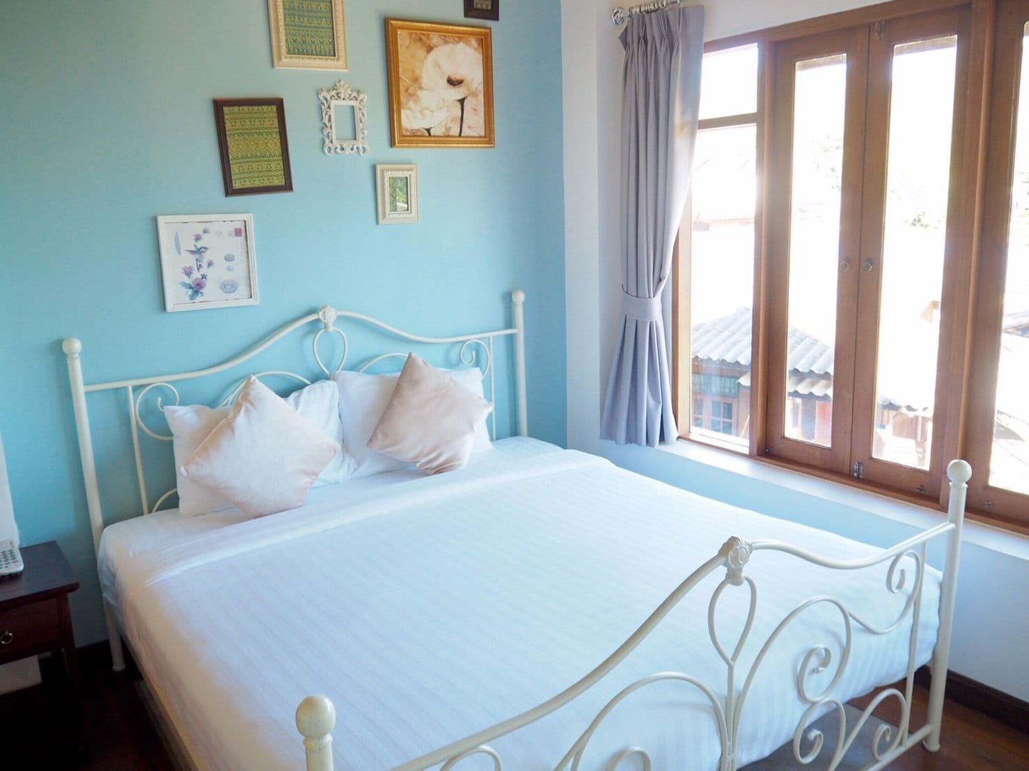 Una cama bien hecha con ropa blanca y estructura metálica, una mesita de noche con un control remoto y una ventana con cortinas.