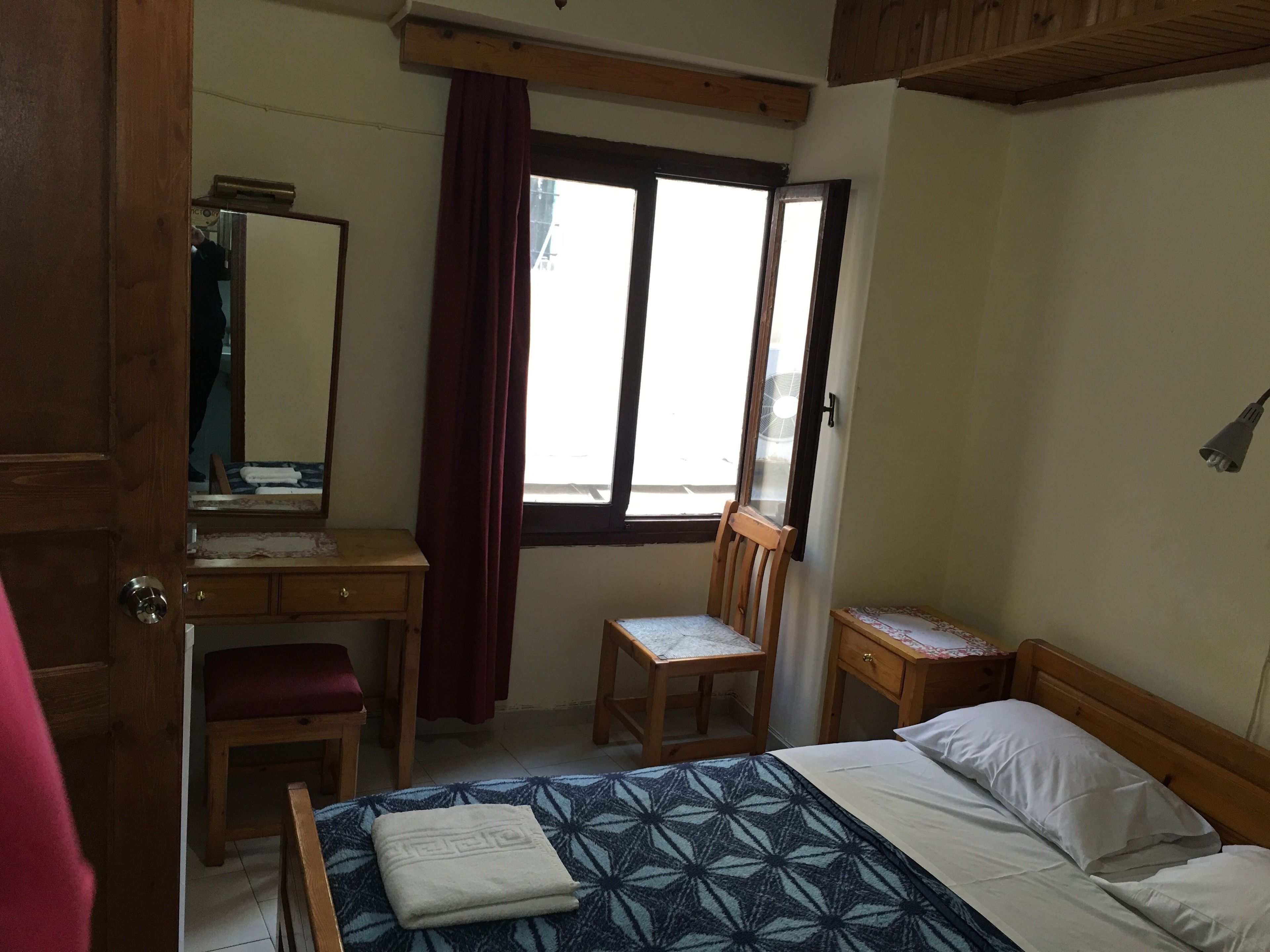 A bedroom with a bed, a wooden desk, a chair, a bedside table, and a window with curtains.