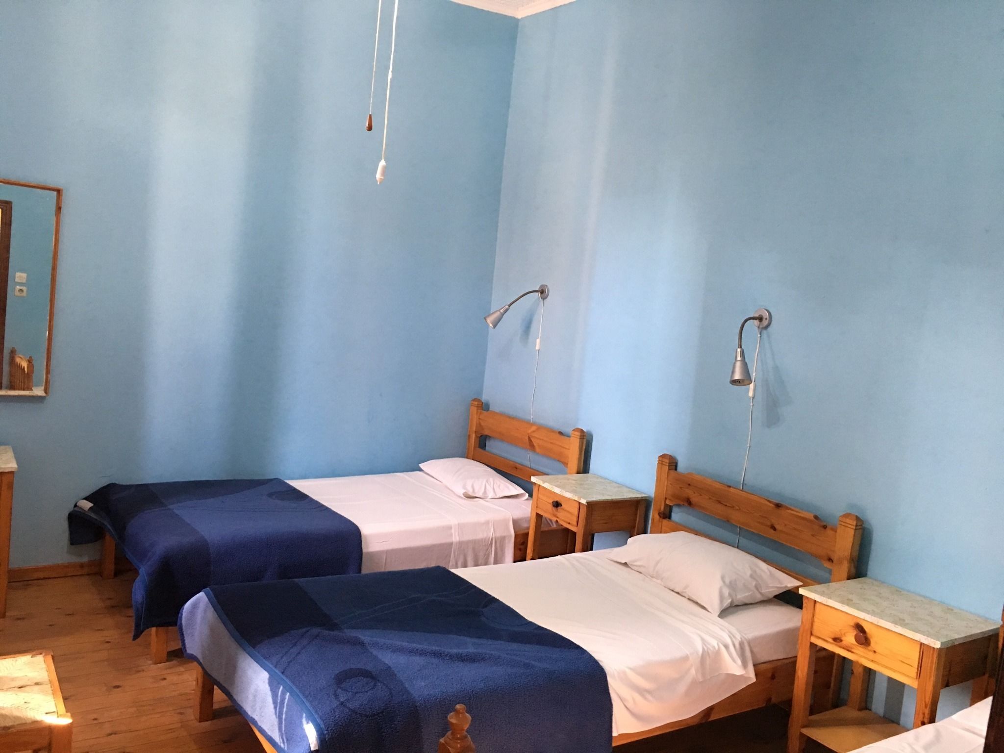 Una habitación con dos camas individuales, cada una con cabecero y mesitas de noche de madera. Hay dos lámparas de pared y un pequeño mueble auxiliar de madera con un cajón.