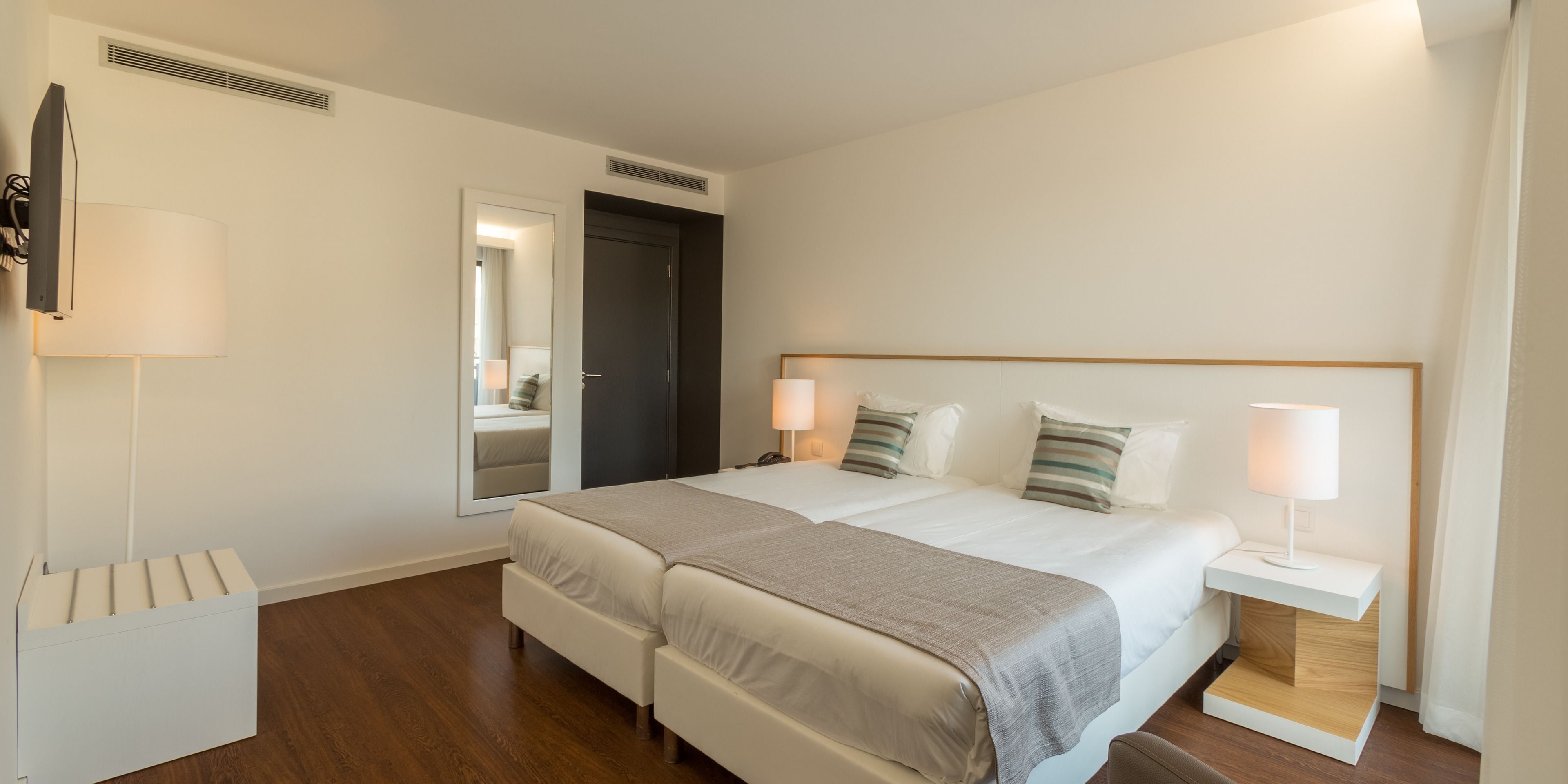 Une chambre d’hôtel moderne avec deux lits, une table de chevet avec une lampe, un miroir et une armoire.