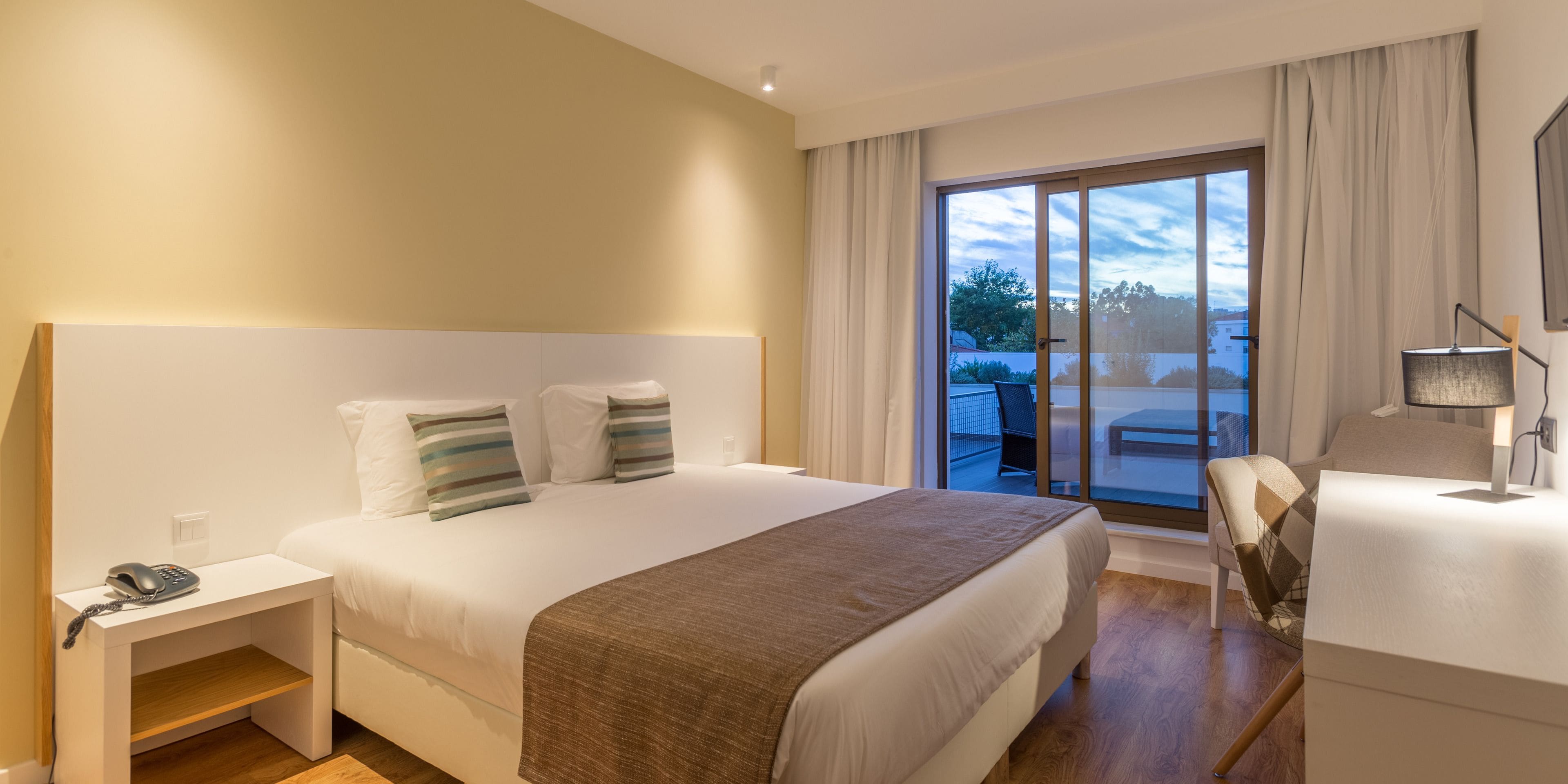 Habitación de hotel con una cama grande, una mesita de noche con teléfono, una silla, un escritorio con lámpara y vistas al exterior a través de una puerta corrediza de vidrio.
