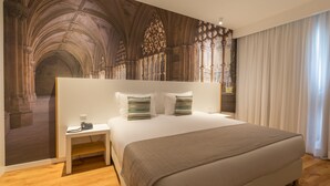 A hotel room with a large bed, a nightstand, a telephone, and a view of a cathedral interior through a large window. - Steyler Fatima Hotel Congress (Ourem)