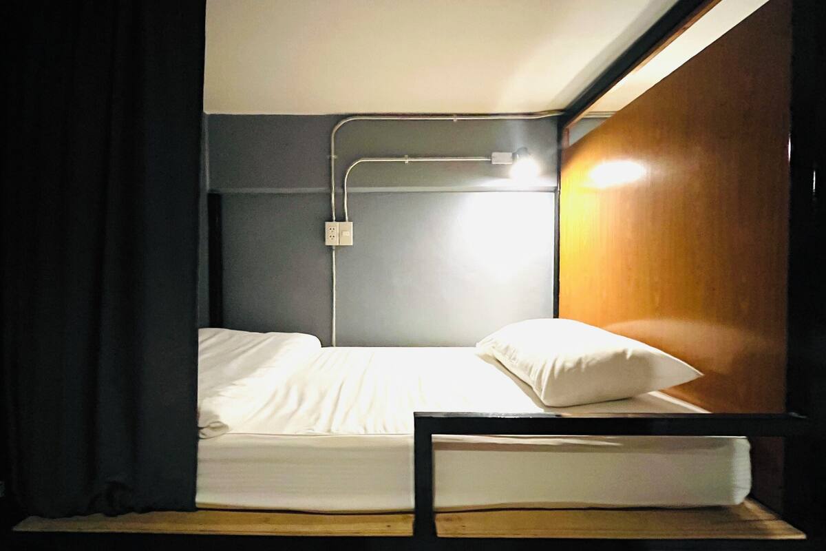 A small, single-bed room with a wooden headboard, a bedside table, and a wall-mounted light fixture.