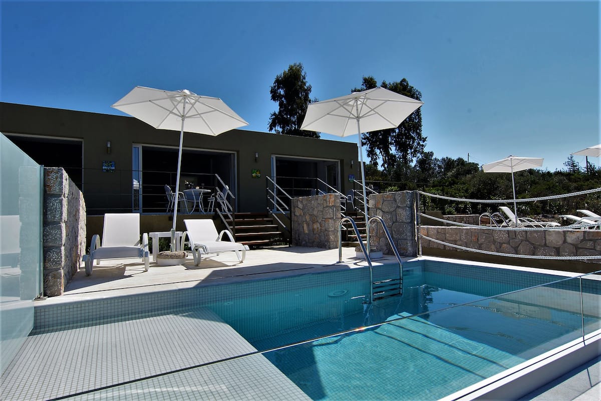 A clear blue pool with white lounge chairs and umbrellas, stone walls, and a staircase leading to a building with a door.