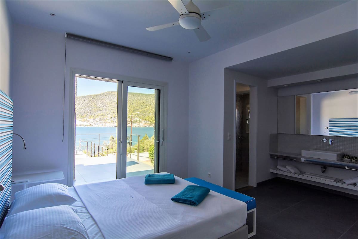 A hotel room with a bed, a balcony with a view, a bathroom with a large mirror, and a ceiling fan.