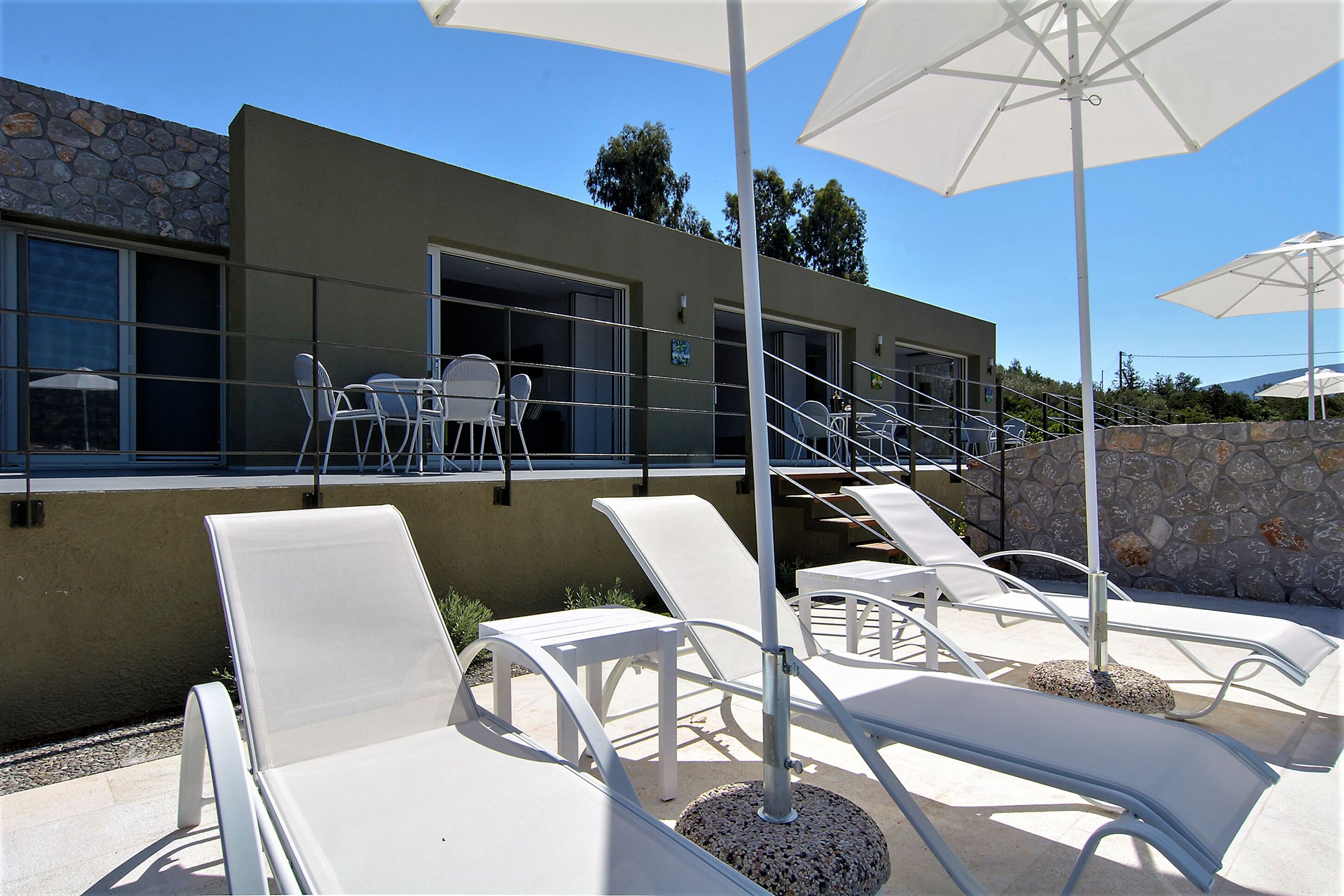 A modern building with a stone facade, outdoor seating area with white lounge chairs and umbrellas, and a staircase leading to an upper level.
