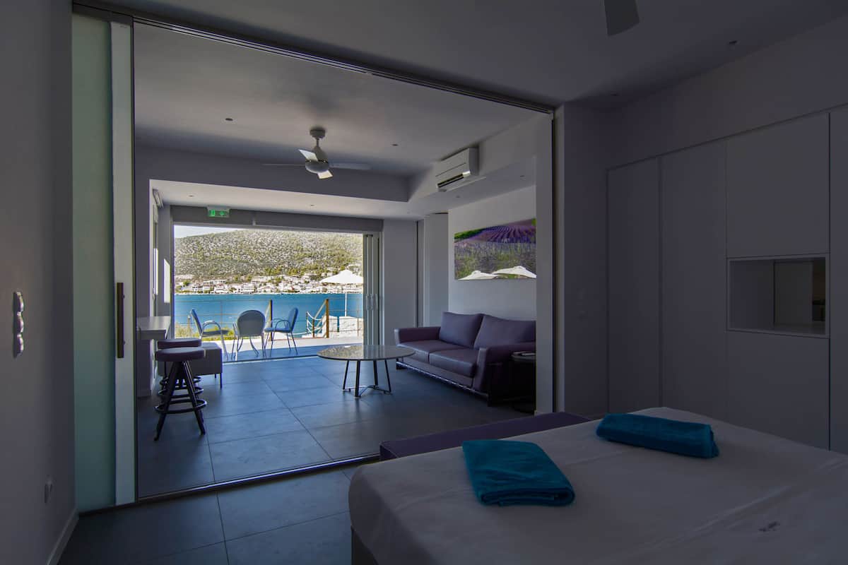 A modern hotel room with a bed, a sofa, a small table, and a balcony with a view of the sea.