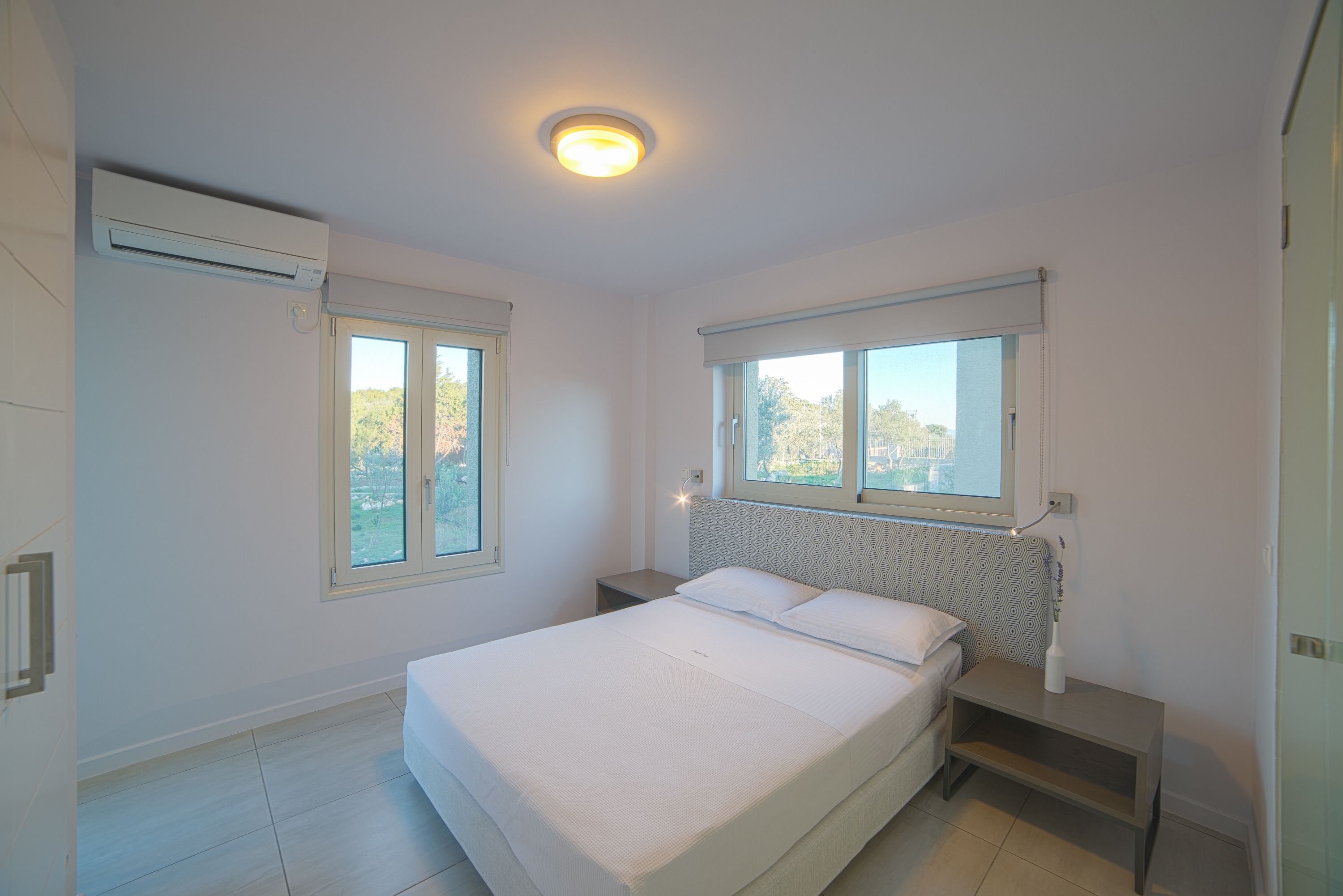 A bedroom with a bed, two bedside tables, a nightstand, a window, and an air conditioning unit.