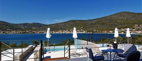 Una terraza con piscina, sombrillas blancas y vistas a un pueblo costero y colinas.