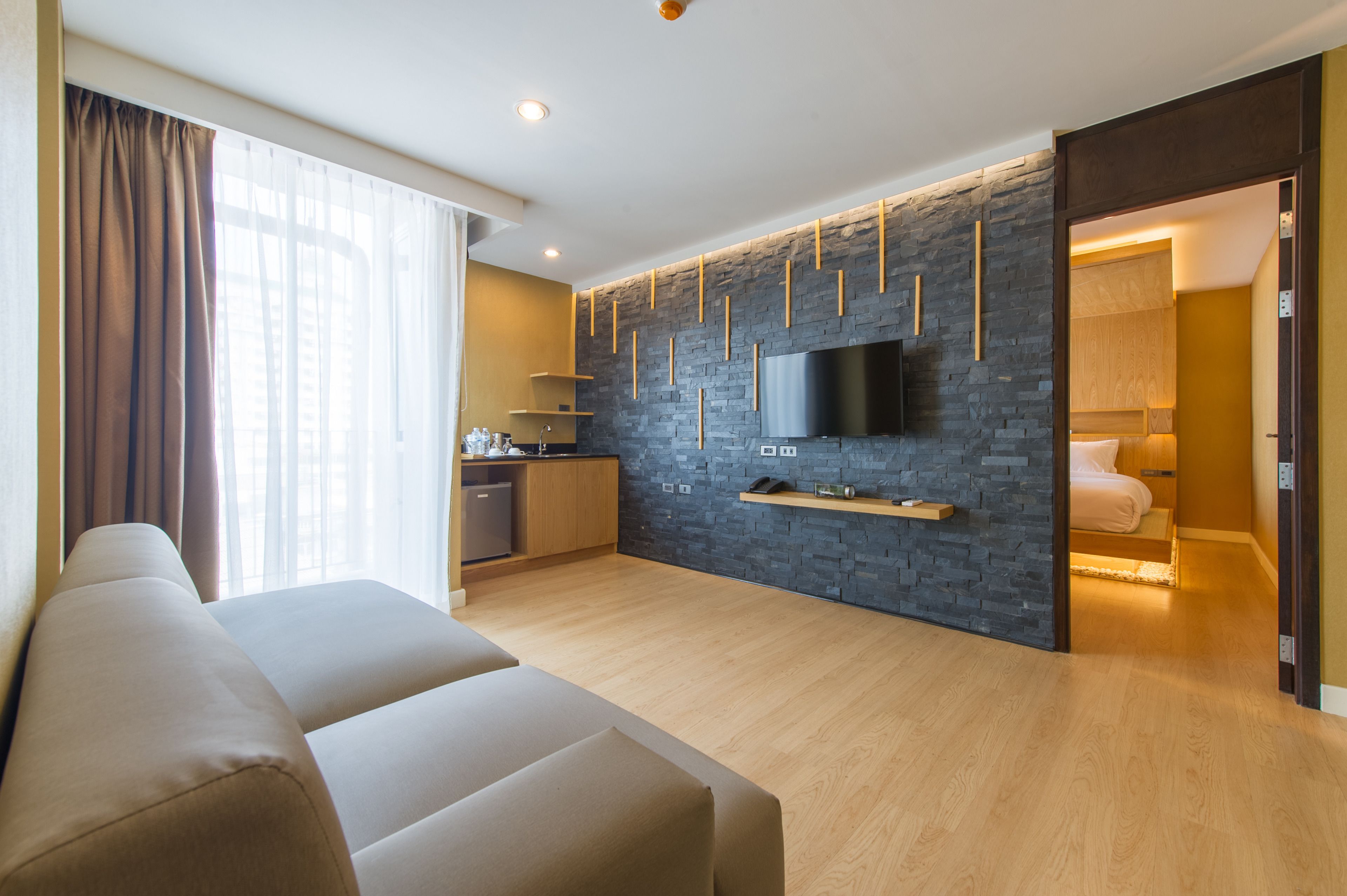 Un saló modern amb un sofà, una zona de cuina amb rentaplats, un televisor de pantalla plana fixat a una paret de pedra i un dormitori amb llit i tauleta de nit.