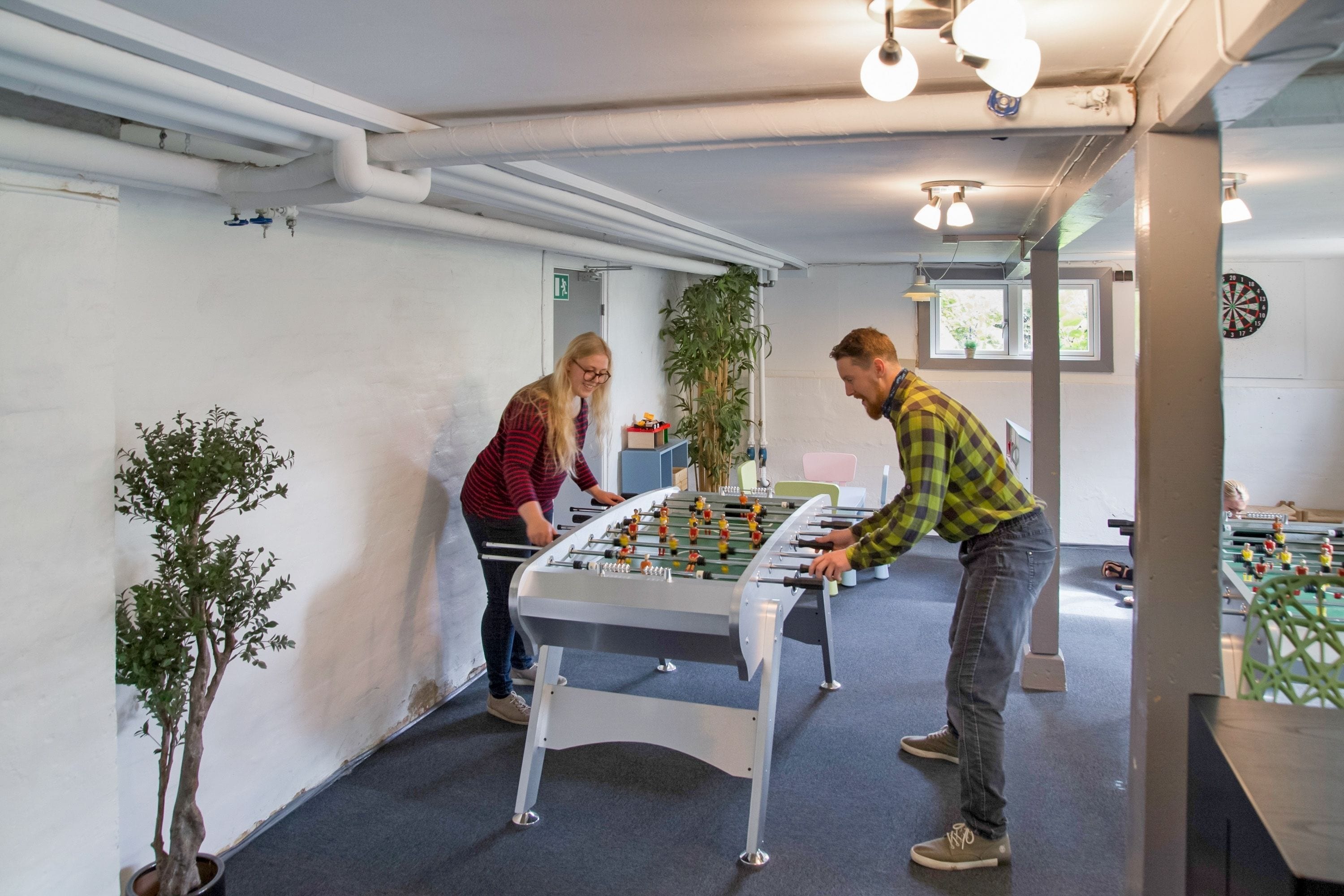 A room with a foosball table, a dartboard, and a foosball table in the background.