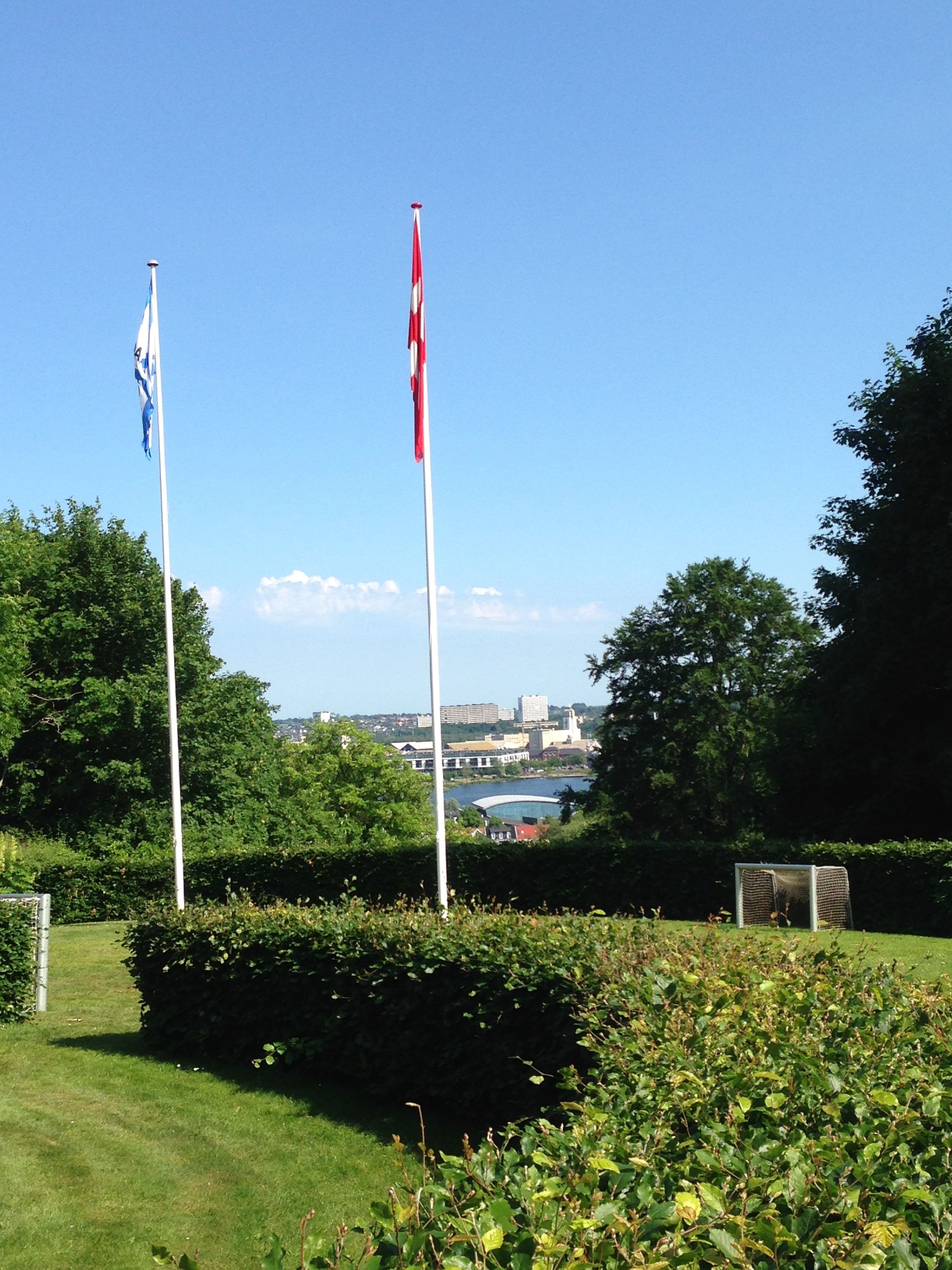 Deux mâts surmontés de drapeaux, une zone herbeuse et une vue lointaine sur des bâtiments et une étendue d’eau.