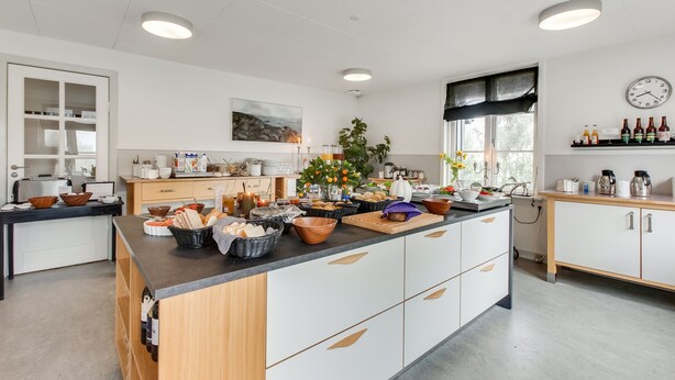 A modern kitchen with a central island, white cabinetry, and a variety of food items and kitchenware. - Danhostel Kolding (Kolding)