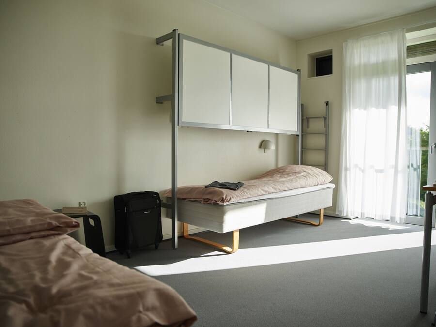 A bedroom with a bunk bed, a desk, a chair, a suitcase, and a window with curtains.