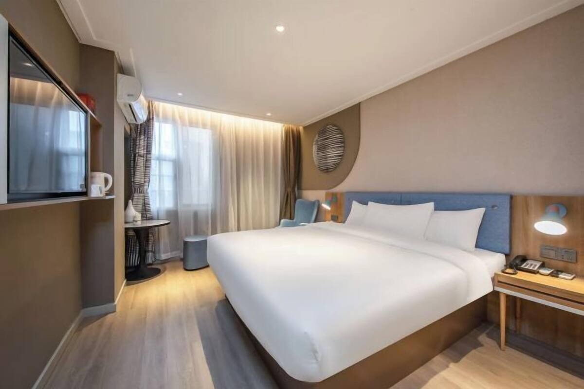 A modern hotel room with a large bed, a bedside table with a lamp, a small desk, and a television.