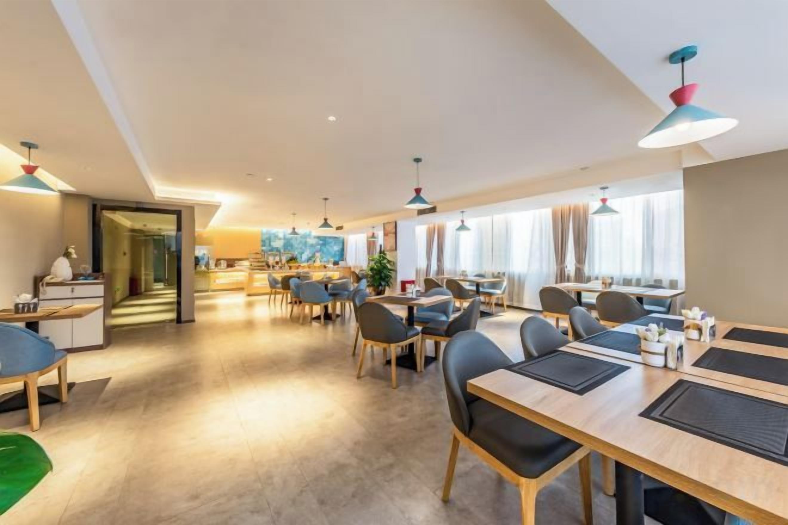 A modern hotel dining area with multiple tables and chairs, a kitchen in the background, and large windows allowing natural light.
