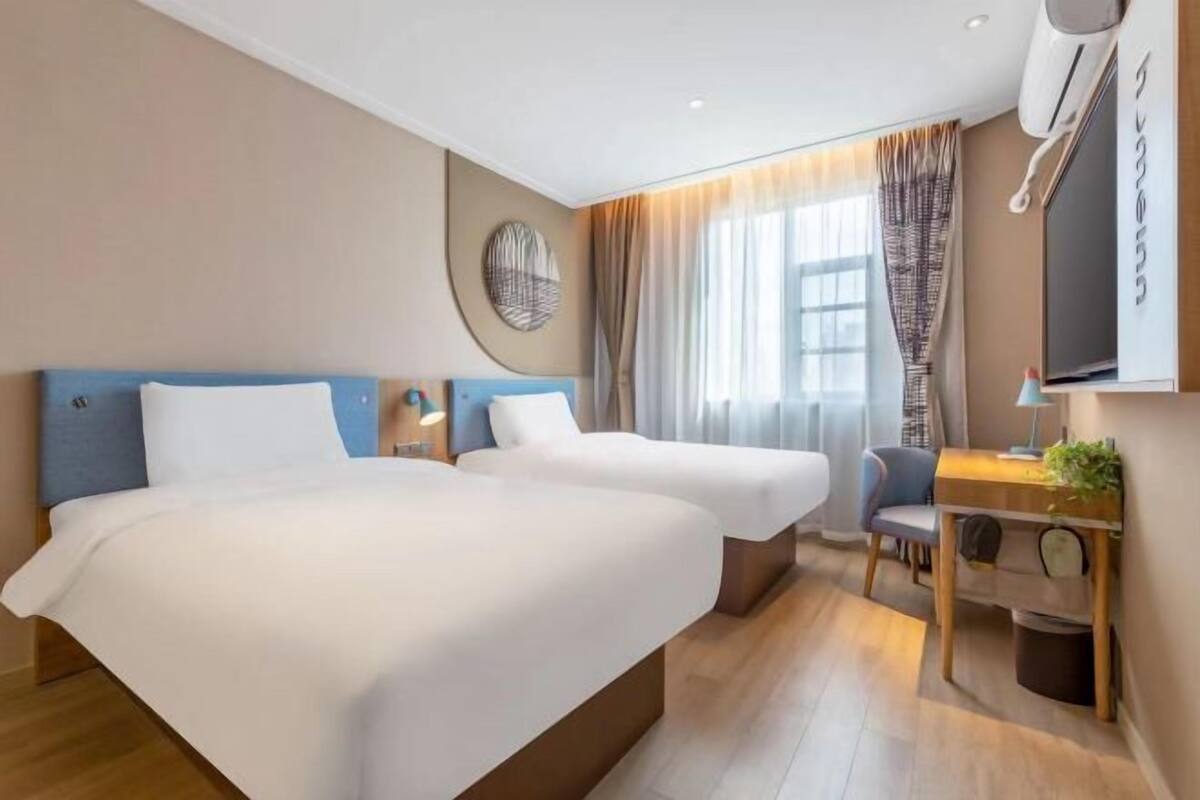 A hotel room with two beds, a desk, a chair, a TV, and a window with curtains.