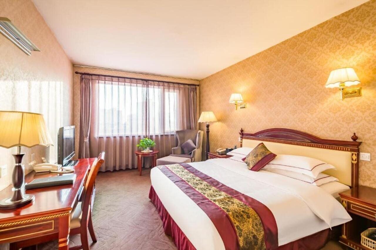 A hotel room with a large bed, a desk with a lamp, a chair, a television, and a window with curtains.