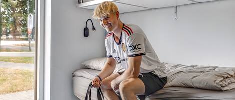 A person sitting on a bed in a room with a bunk bed above, a window, and a door.