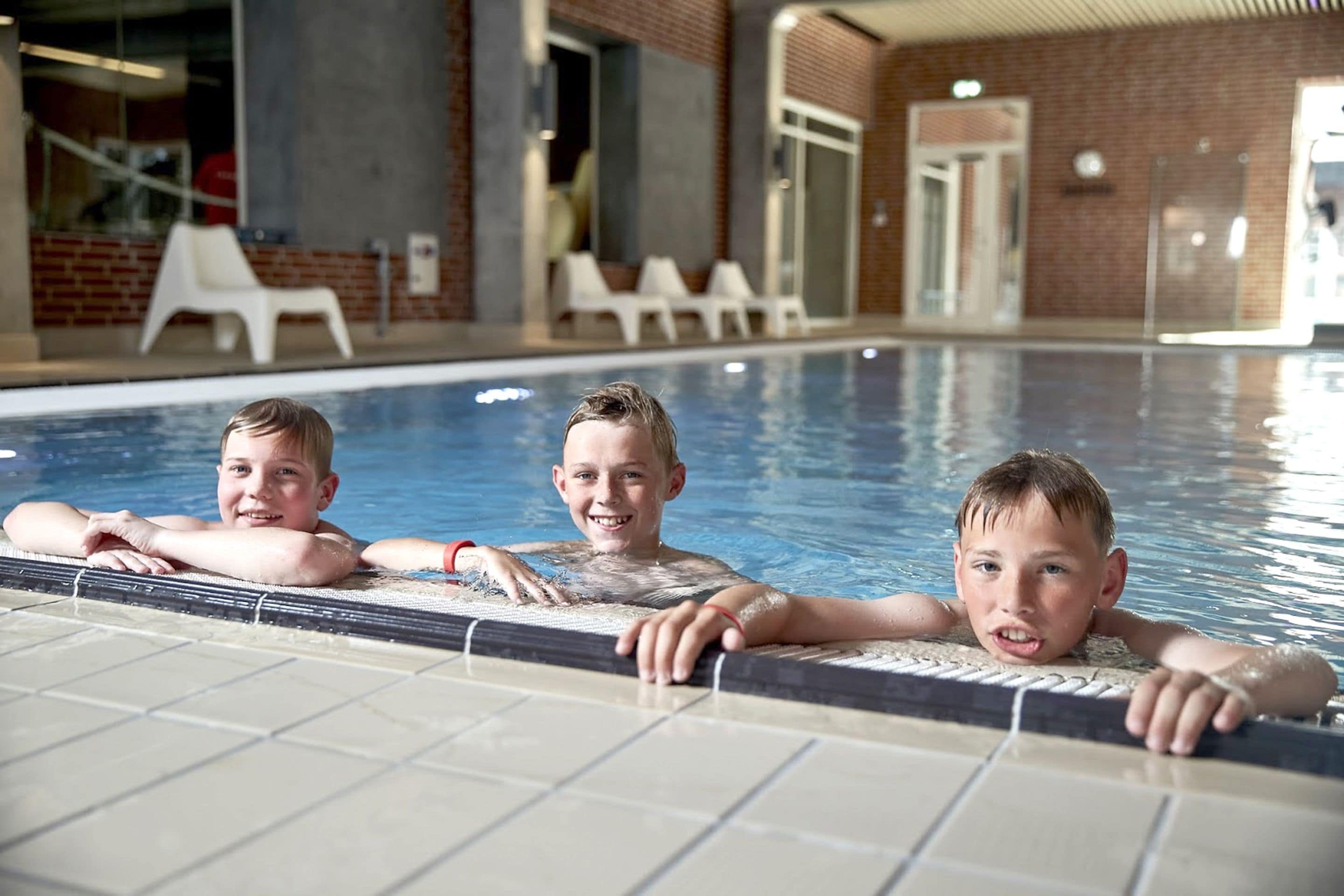 Una piscina interior con agua cristalina, suelo de baldosas y sillones blancos al fondo.