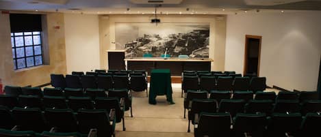Konferenssisali, jossa on riveissä järjestettyjä tuoleja, edessä puhujapönttö, seinällä suuri mustavalkoinen seinämaalaus ja ikkuna, josta avautuu näkymä rakennuksiin.