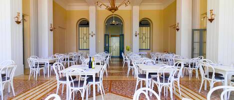 A spacious dining area with white tables and chairs, a patterned floor, and a central chandelier.