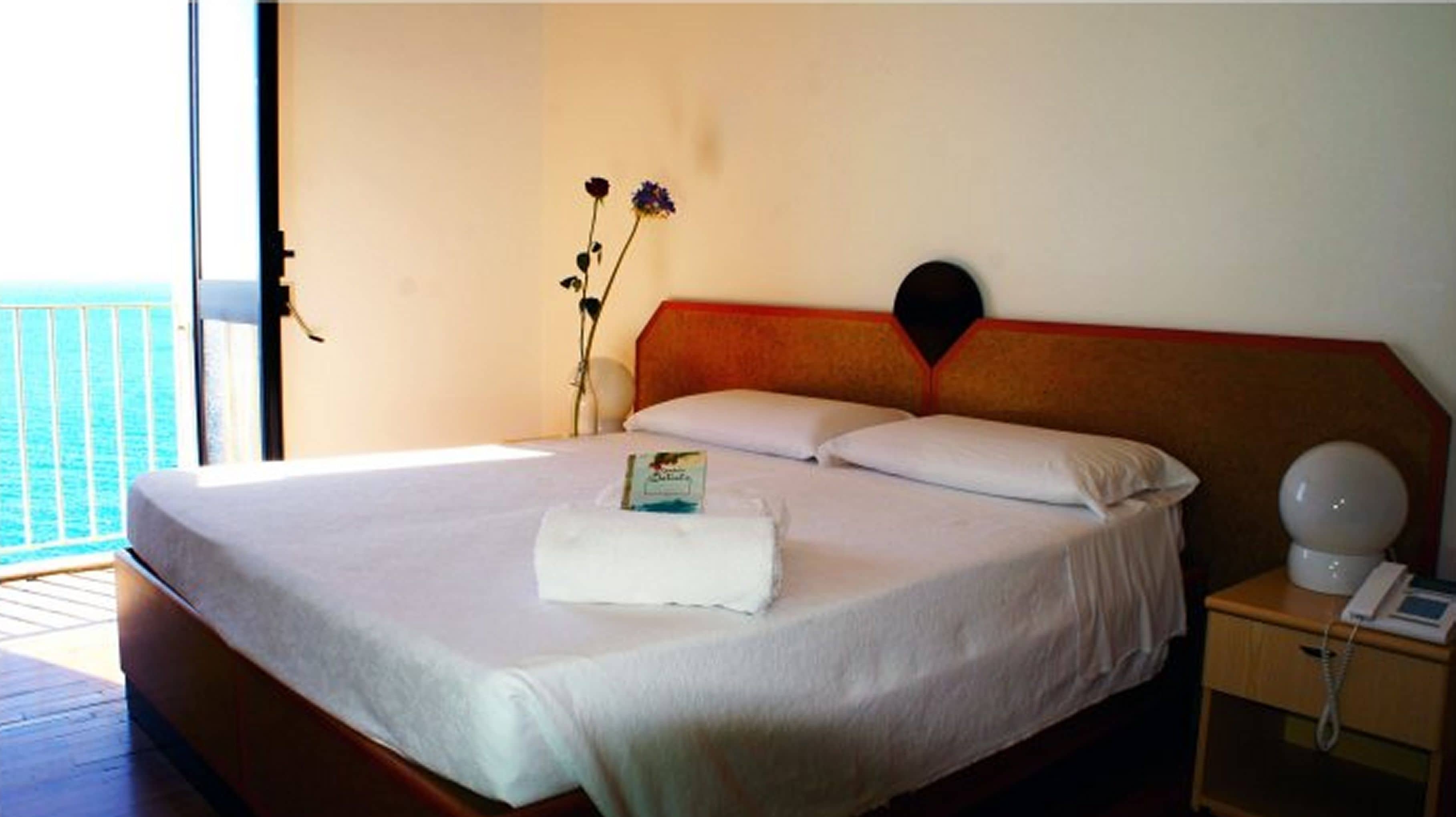 Een hotelkamer met een bed, een nachtkastje met een lamp en uitzicht op zee.