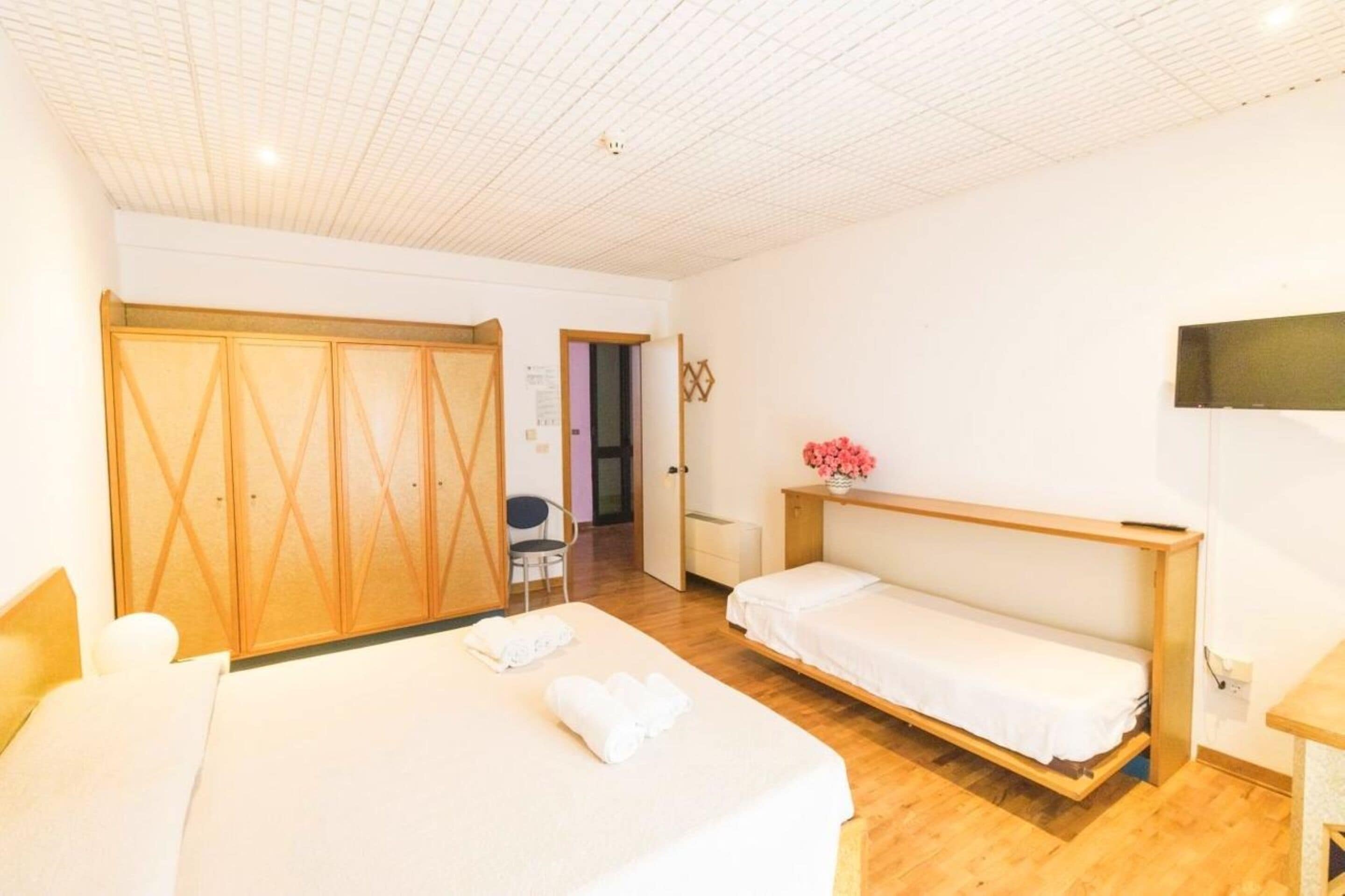 A hotel room with a large bed, a smaller bed, a wooden wardrobe, a chair, and a small table with a vase of flowers.