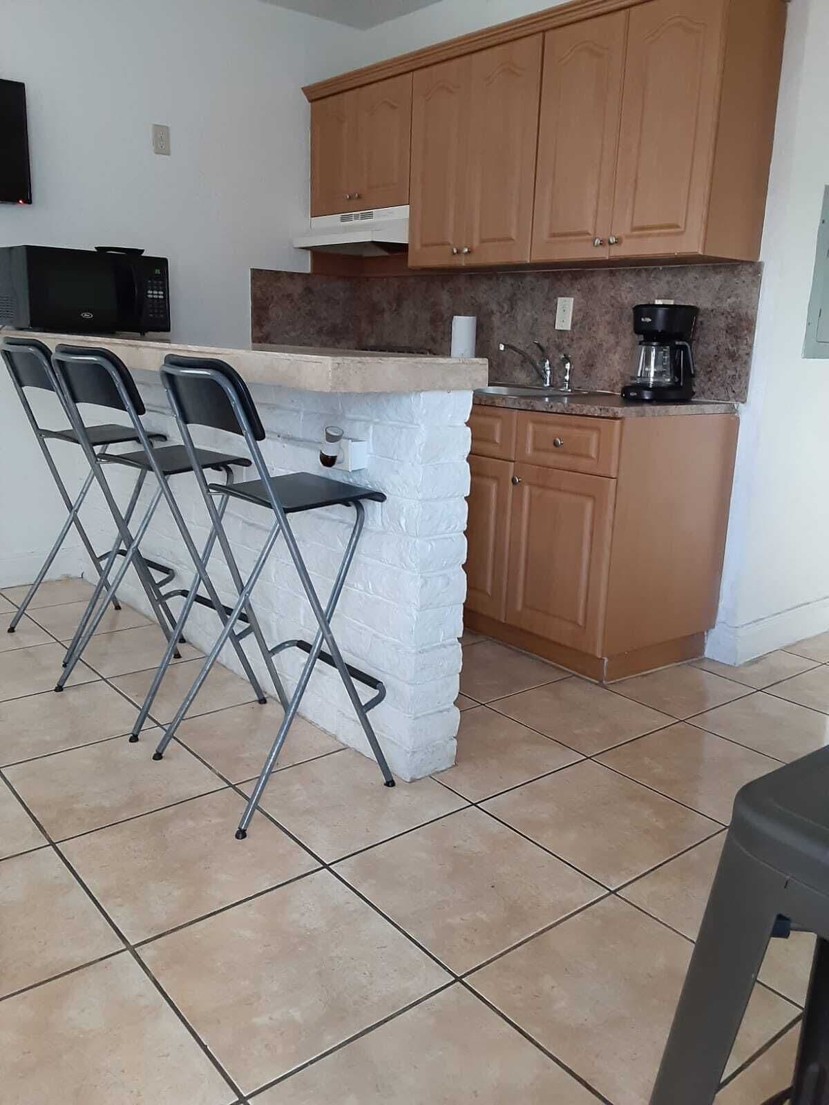 A compact kitchen with wooden cabinets, a white countertop, and a coffee maker. There are three foldable chairs against the wall.