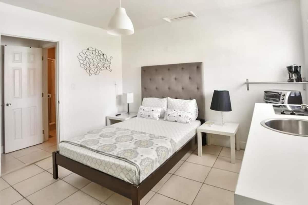 Una camera da letto moderna con un letto, comodini, una lampada e un angolo cottura integrato con lavandino.