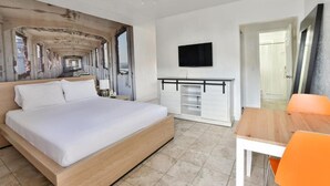 Ein Zimmer mit einem Bett, einem an der Wand montierten Fernseher, einem Schreibtisch mit Stuhl und einer Tür zu einem weiteren Raum.