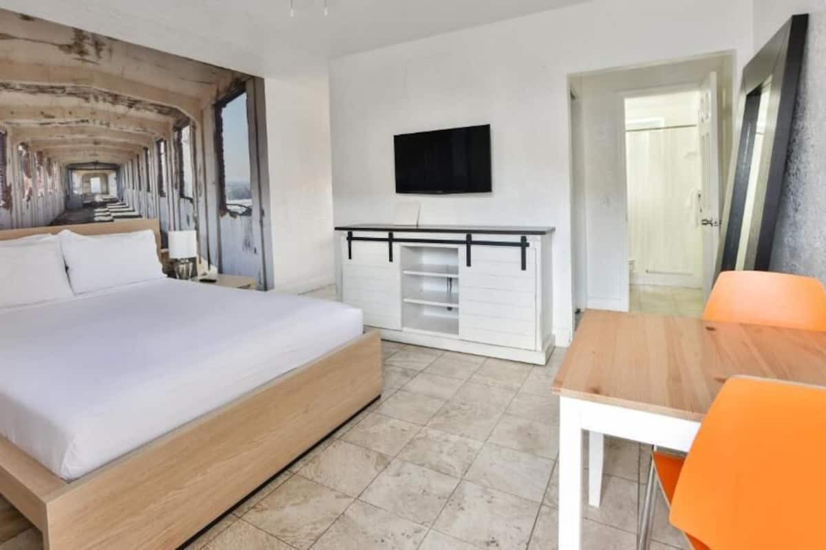 Una camera con un letto, una TV fissata al muro, una scrivania con una sedia e una porta che conduce a un'altra stanza.
