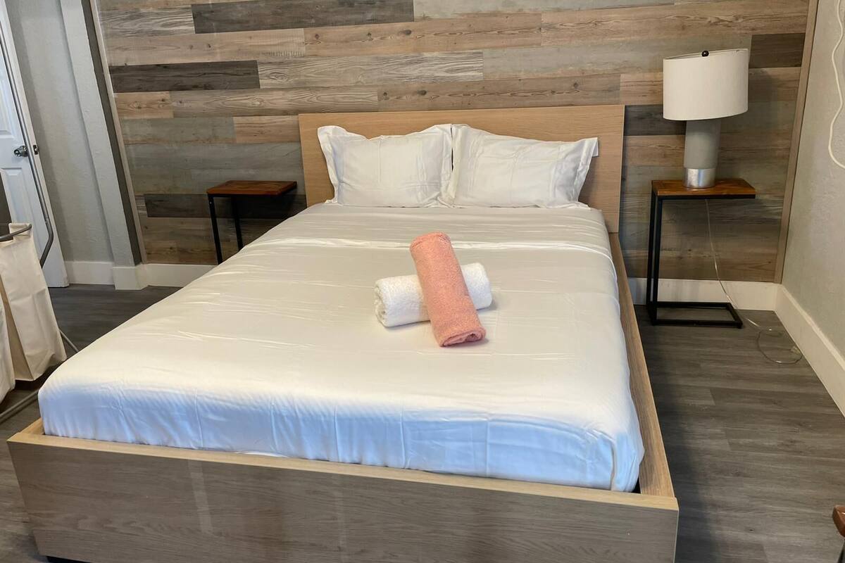 Un letto rifatto con lenzuola bianche e un asciugamano rosa, una testiera in legno, un comodino con una lampada e un pavimento di legno.