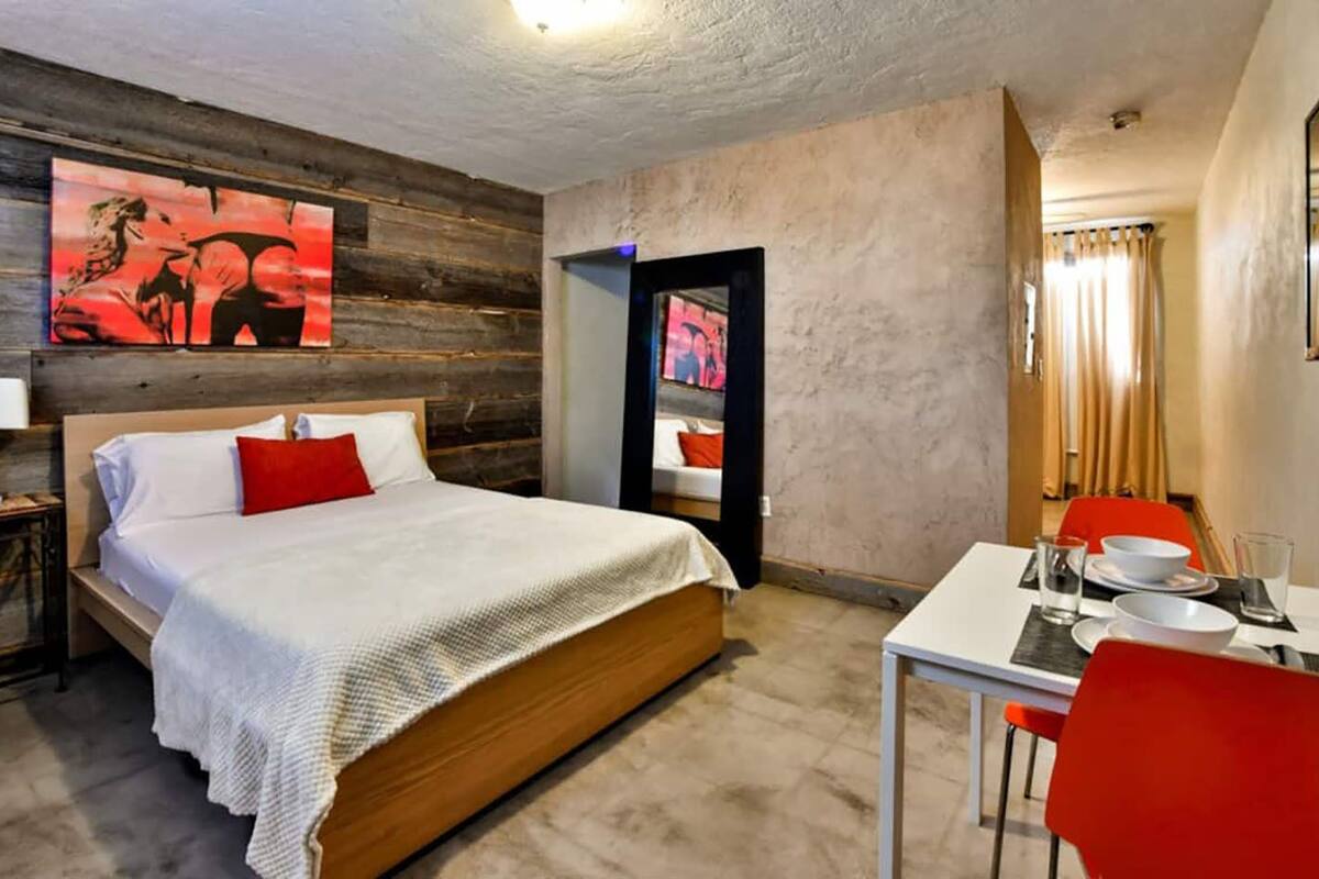 Una camera d'albergo con una testiera in legno, un letto con biancheria da letto bianca e un cuscino rosso, un tavolo da pranzo con sedie rosse e opere d'arte alle pareti.