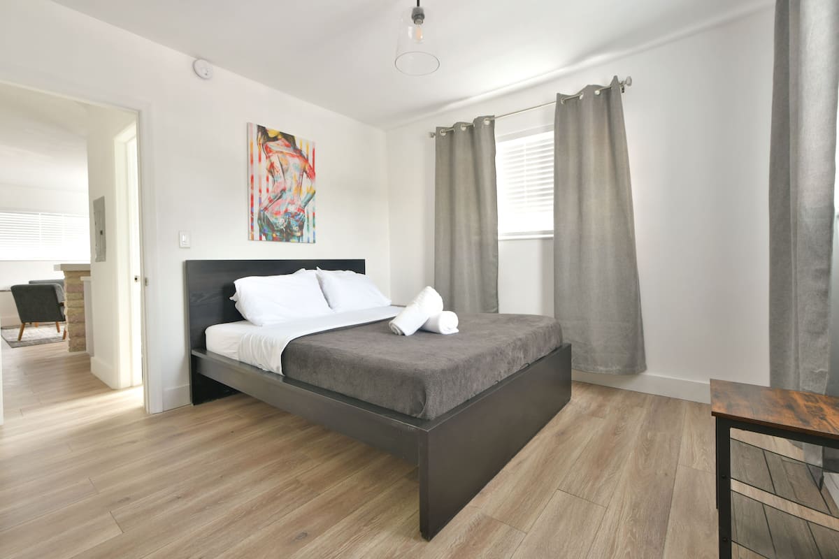 Una camera da letto moderna con un letto grande, pavimento in legno, un quadro appeso e vista all'esterno tramite una finestra con tende.