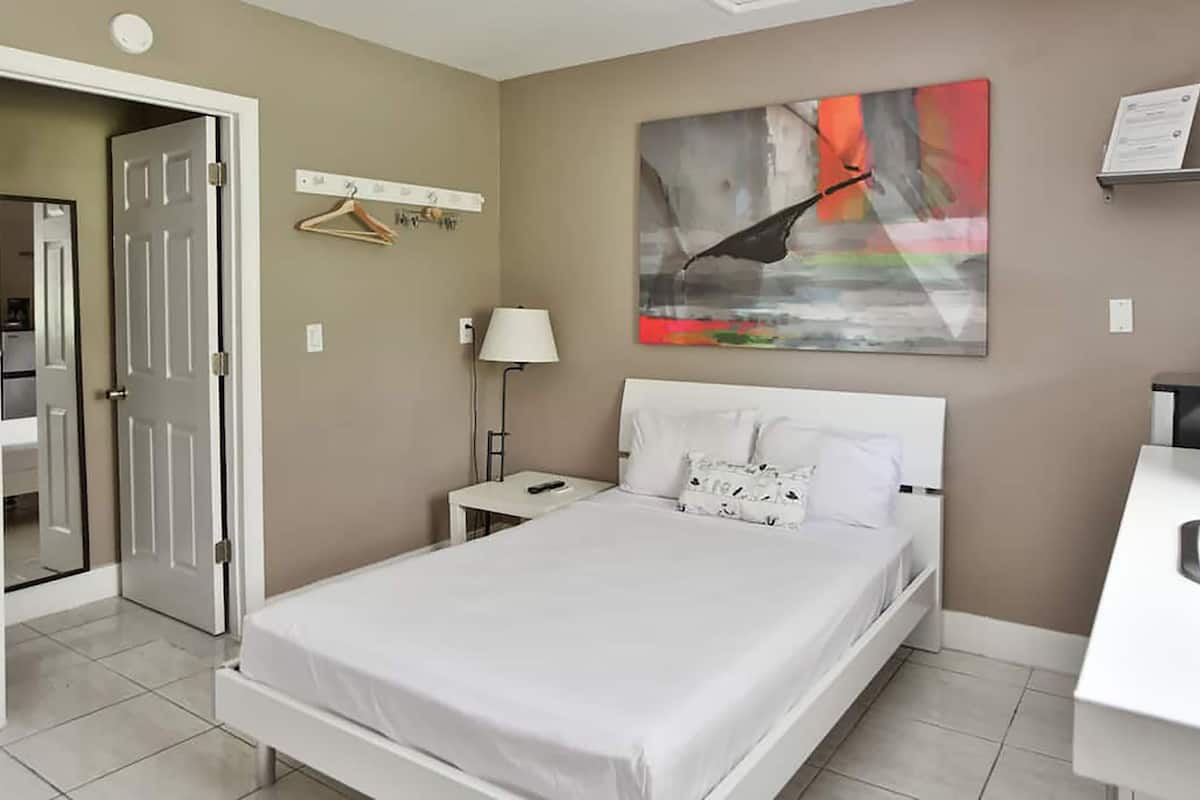 Una camera d'albergo con un letto, un comodino con una lampada, un quadro appeso alla parete e un angolo cottura.