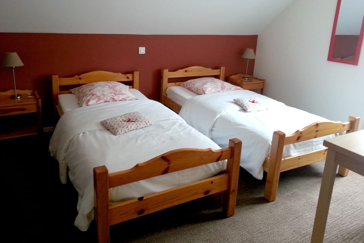Two wooden beds with white bedding and floral pillows, a wooden nightstand with a lamp, and a small table in a room with red walls.