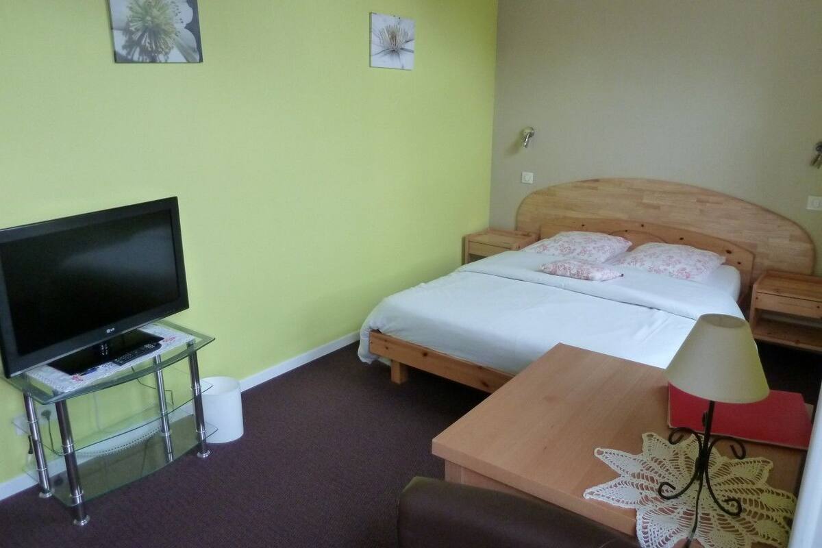 A room with a bed, a TV on a glass stand, a wooden table, and a lamp.