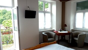 Un dormitorio con cama, dos sillas, una mesita, televisión y ventanas amplias.